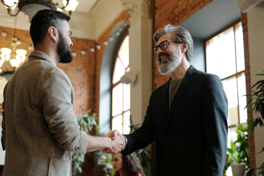 Two businessmen smiling during a handshake in a modern office setting, symbolizing success and partnership.