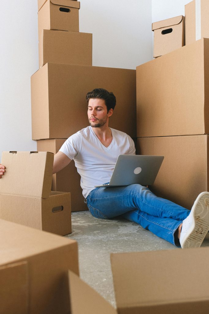 Young focused male freelancer checking items in carton box while sitting on floor and working online on laptop before moving into new flat