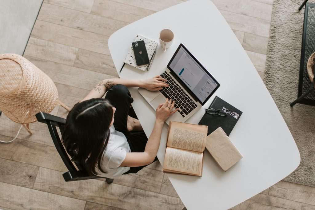 Top view of young woman in casual wear browsing netbook and reading book while working remotely in comfortable modern workspace